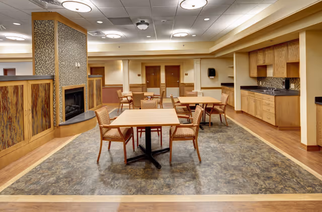Spacious assisted-living dining and common area with several tables and chairs, a fireplace partition, and a kitchenette along one wall.