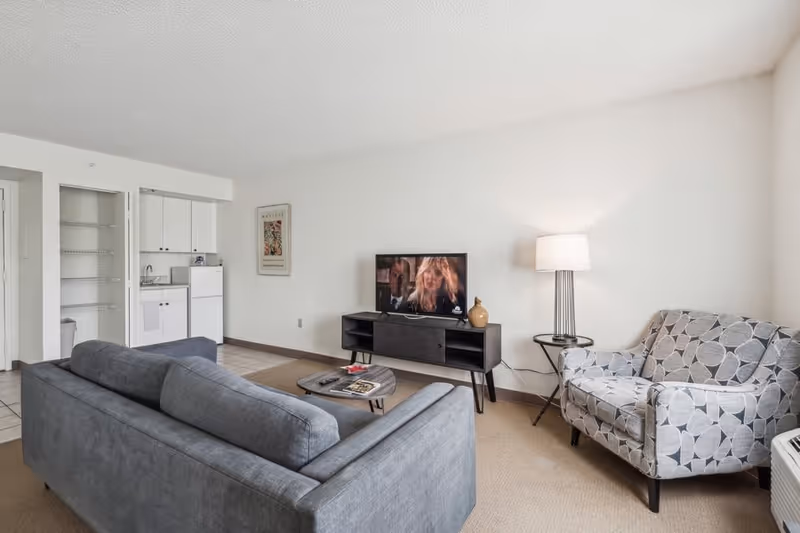 A living room area in a senior living facility featuring a gray sofa, a patterned armchair, a small round coffee table with magazines and a remote control, a TV on a black stand showing a scene from a movie, a floor lamp, and a kitchenette with white cabinets and a small refrigerator in the background.