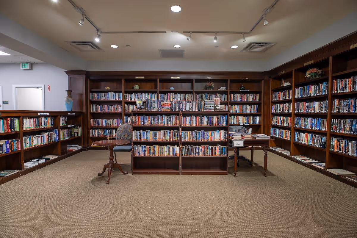 Interior view of a library room with wooden bookshelves filled with books lining the walls and a central bookshelf. Two chairs and two small wooden tables are placed near the shelves. The room has beige carpet flooring and ceiling lights.