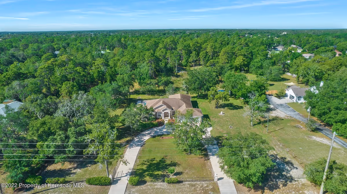 Aerial view of a residential area surrounded by dense green trees and vegetation. A single-story house with a brown roof is centered in the image, with a curved driveway leading to the entrance. The surrounding area includes open grassy spaces and neighboring houses visible in the distance under a clear blue sky.
