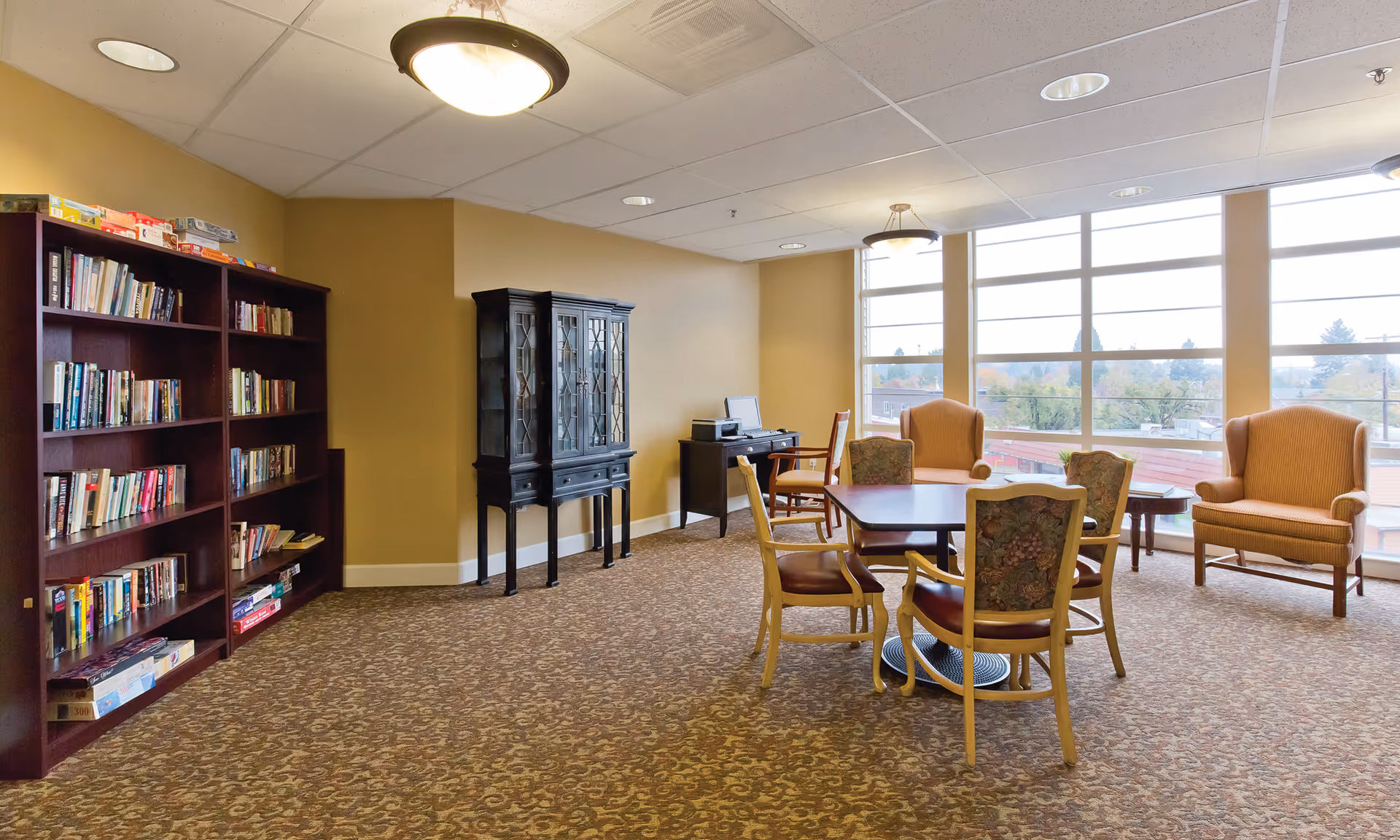A senior living facility common area with large windows letting in natural light. The room features a bookshelf filled with books and board games, a black cabinet, a small desk with a computer and printer, a table with four chairs, and two comfortable armchairs near the windows.