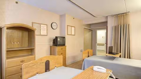 Two wooden beds and furniture in a small shared hospital-style room with a TV.