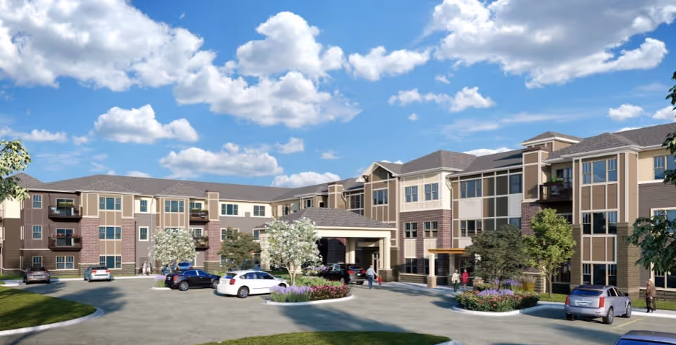 Exterior view of a multi-story senior living facility with a covered entrance, several parked cars, landscaped greenery, and a partly cloudy blue sky.