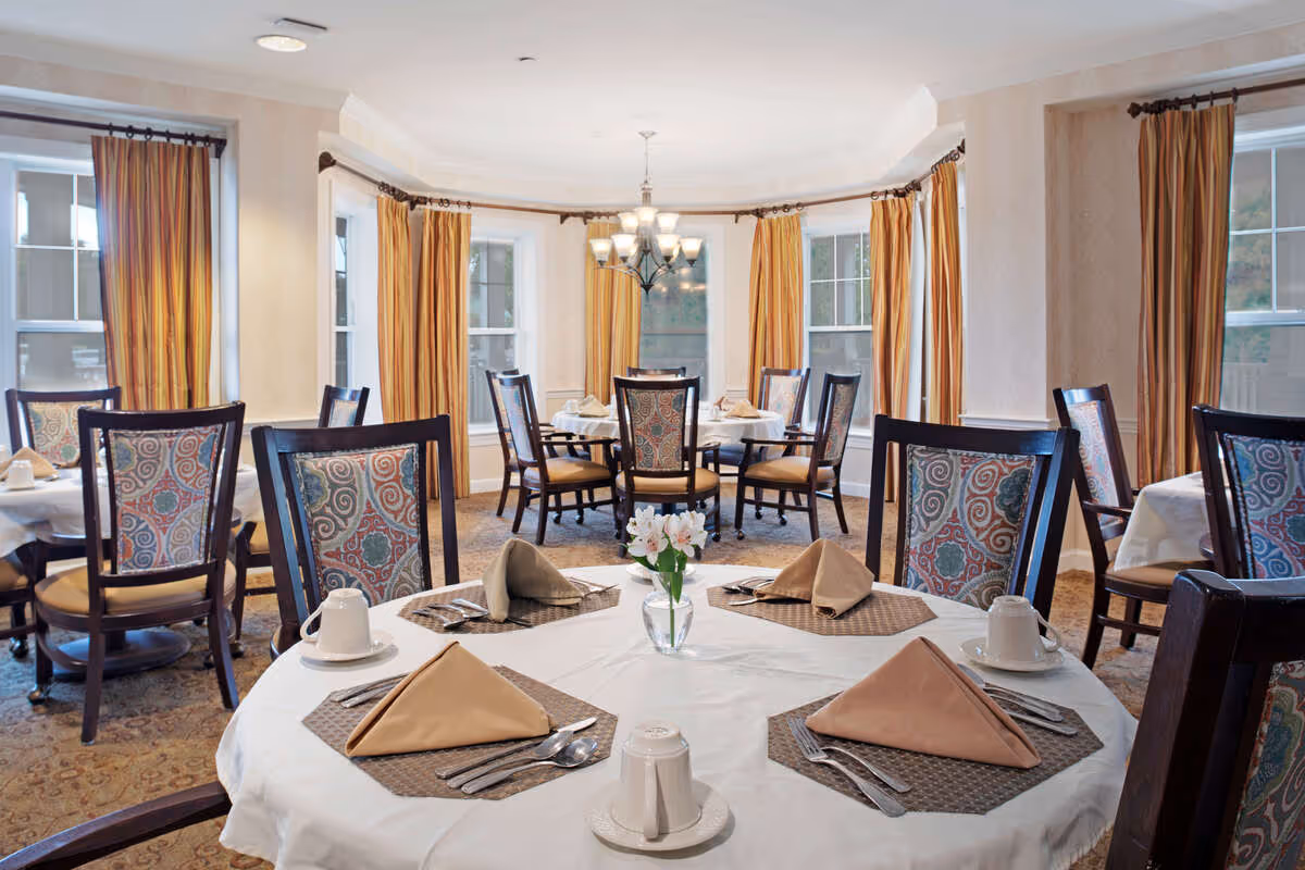 A bright dining room with round tables covered in white tablecloths, each set with beige napkins, silverware, white cups, and a small vase with flowers. The room has multiple windows with yellow and orange striped curtains and a chandelier hanging from the ceiling.