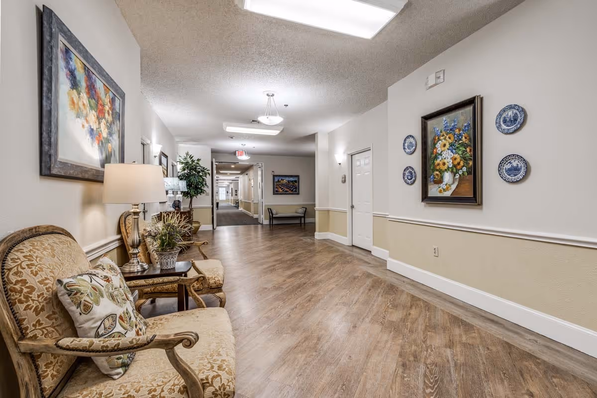 Wide, well-lit interior hallway with cushioned chairs, artwork on the walls, and wood-look flooring.