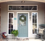Front entrance of a house with a light blue door decorated with a green wreath. The door is flanked by two tall windows with white frames. To the right of the door is a rocking chair with a cushion, and to the left is a small plant stand with a green plant and a decorative cart with flowers. The house number 13701 is displayed vertically next to the door.