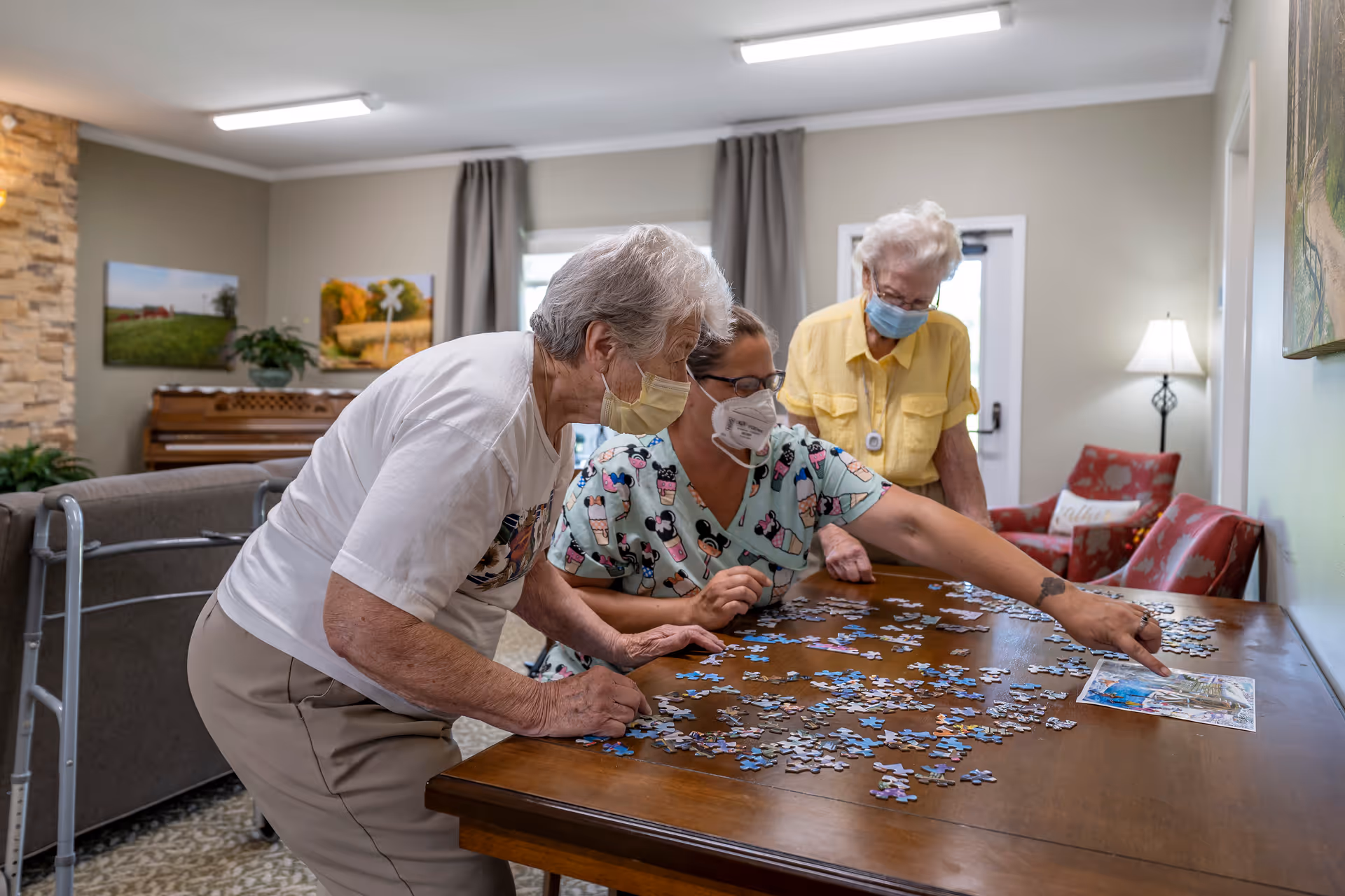 Three elderly women wearing face masks are gathered around a wooden table working on a jigsaw puzzle in a well-lit living room with a piano, paintings, and comfortable chairs in the background.