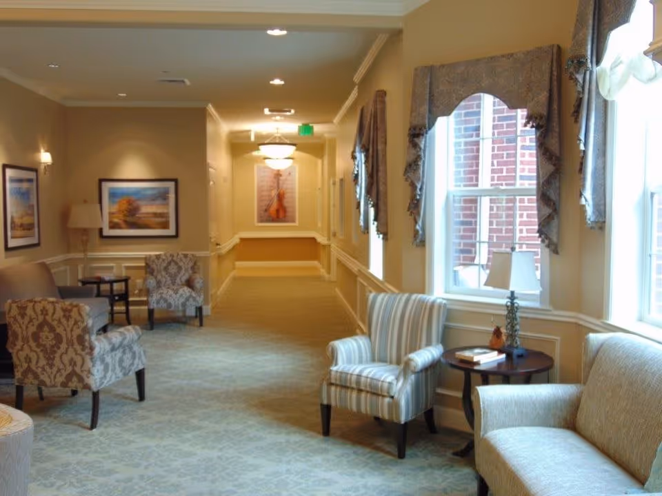A well-lit hallway in a senior living facility with comfortable seating including patterned and striped armchairs, a sofa, side tables with lamps, framed artwork on the walls, and large windows with decorative curtains letting in natural light.