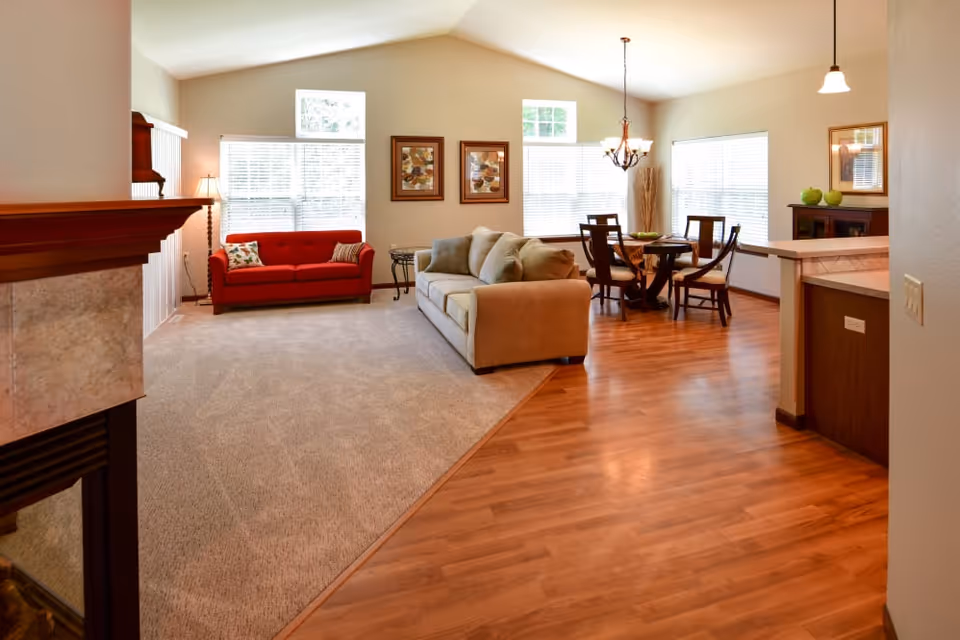 A bright and spacious living and dining area with a beige sofa and a red loveseat on a carpeted section. The dining area features a round wooden table with four chairs on a wooden floor. Large windows with blinds allow natural light to fill the room. A fireplace is partially visible on the left side, and decorative framed artwork hangs on the wall.