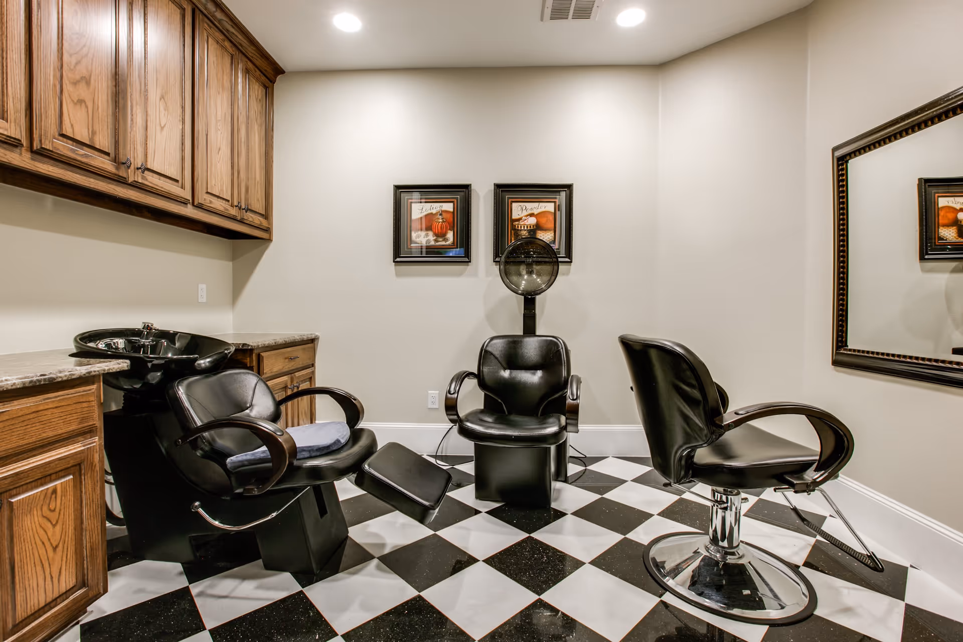 Small salon-style room with three black styling chairs, a hair-washing sink and hood dryer, wooden cabinets, and a large mirror over a black-and-white checkered floor.