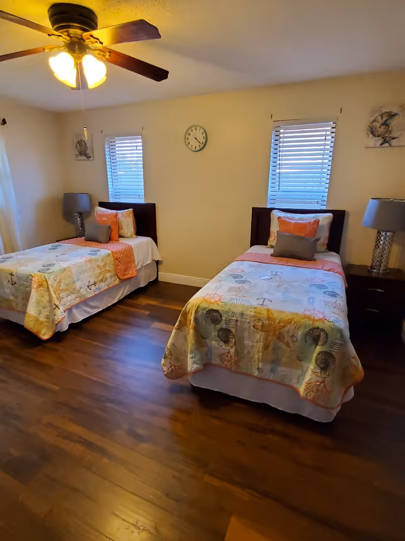 A bedroom with two twin beds, each with colorful seashell and starfish patterned bedspreads and multiple pillows. There are two windows with blinds on the wall behind the beds, a clock centered between the windows, and two bedside tables with lamps. The room has wooden flooring and a ceiling fan with lights.