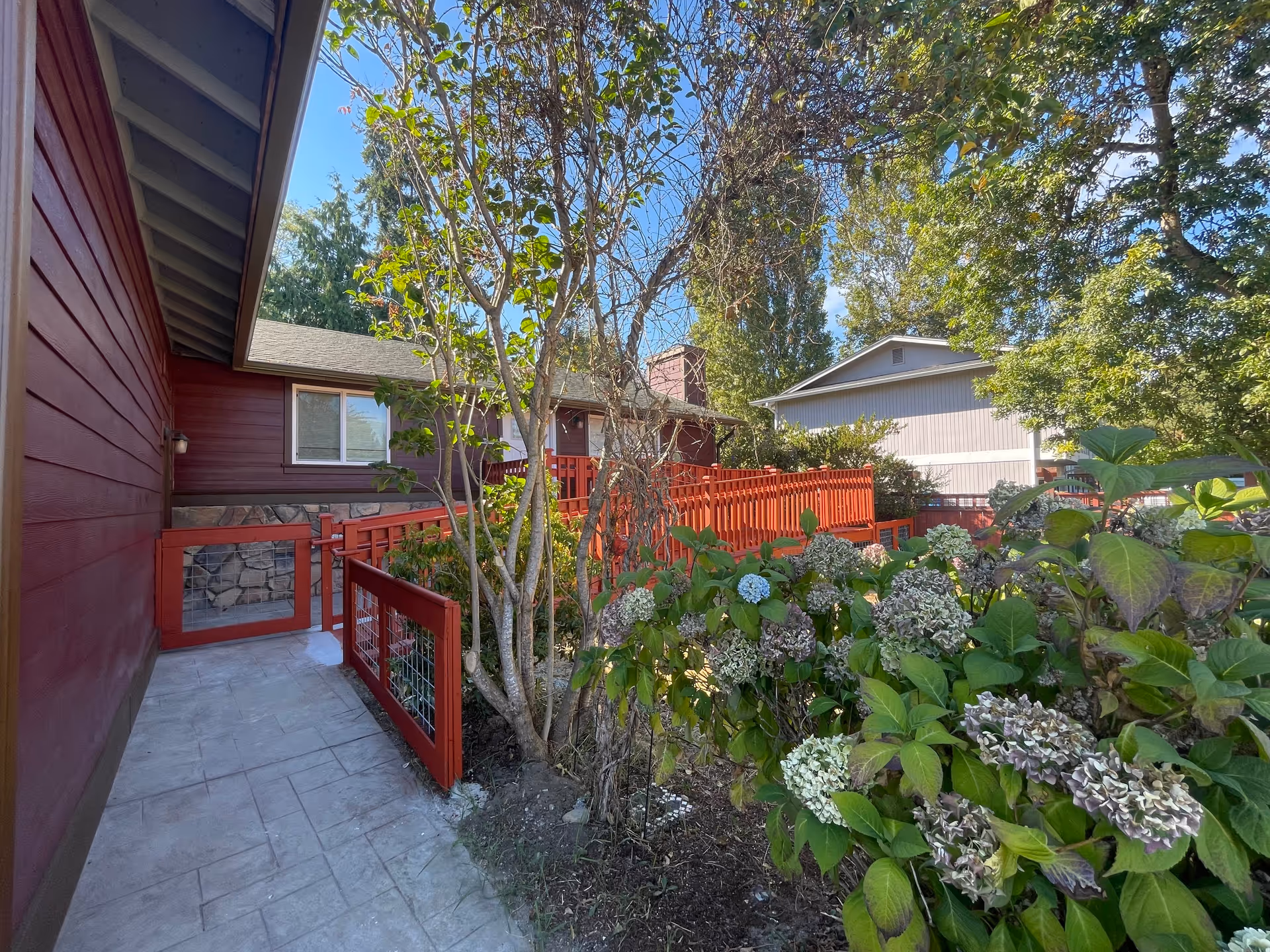 Outdoor view of a red-painted building with a stone foundation, featuring a concrete pathway and a red wooden railing. There are trees and flowering bushes with green leaves and some hydrangea flowers in the foreground under a clear blue sky.