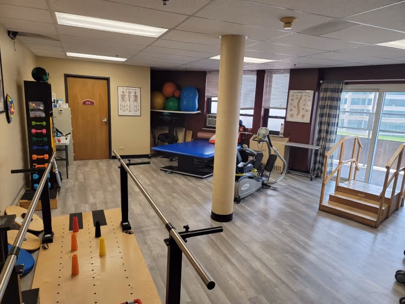 A rehabilitation therapy room with parallel bars for walking exercises, a stationary exercise bike, a blue therapy table, various exercise balls on shelves, anatomical charts on the walls, and a small wooden staircase near large windows with blinds and curtains.