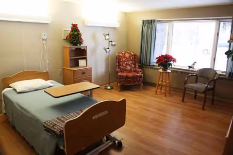 Interior hospital-style patient room with a bed, nightstand, chair, and large window.