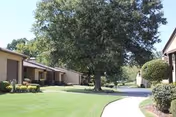 A paved walkway curves through a well-maintained outdoor area with green grass, bushes, and trees. Single-story buildings with beige walls and brown roofs line the walkway on both sides under a clear sky.