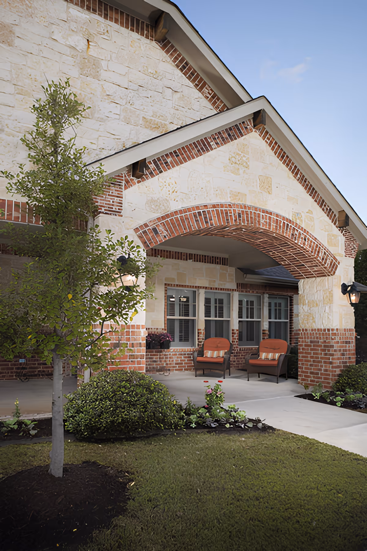 Stone-and-brick front entrance porch with two cushioned chairs, potted plants, and landscaped lawn.