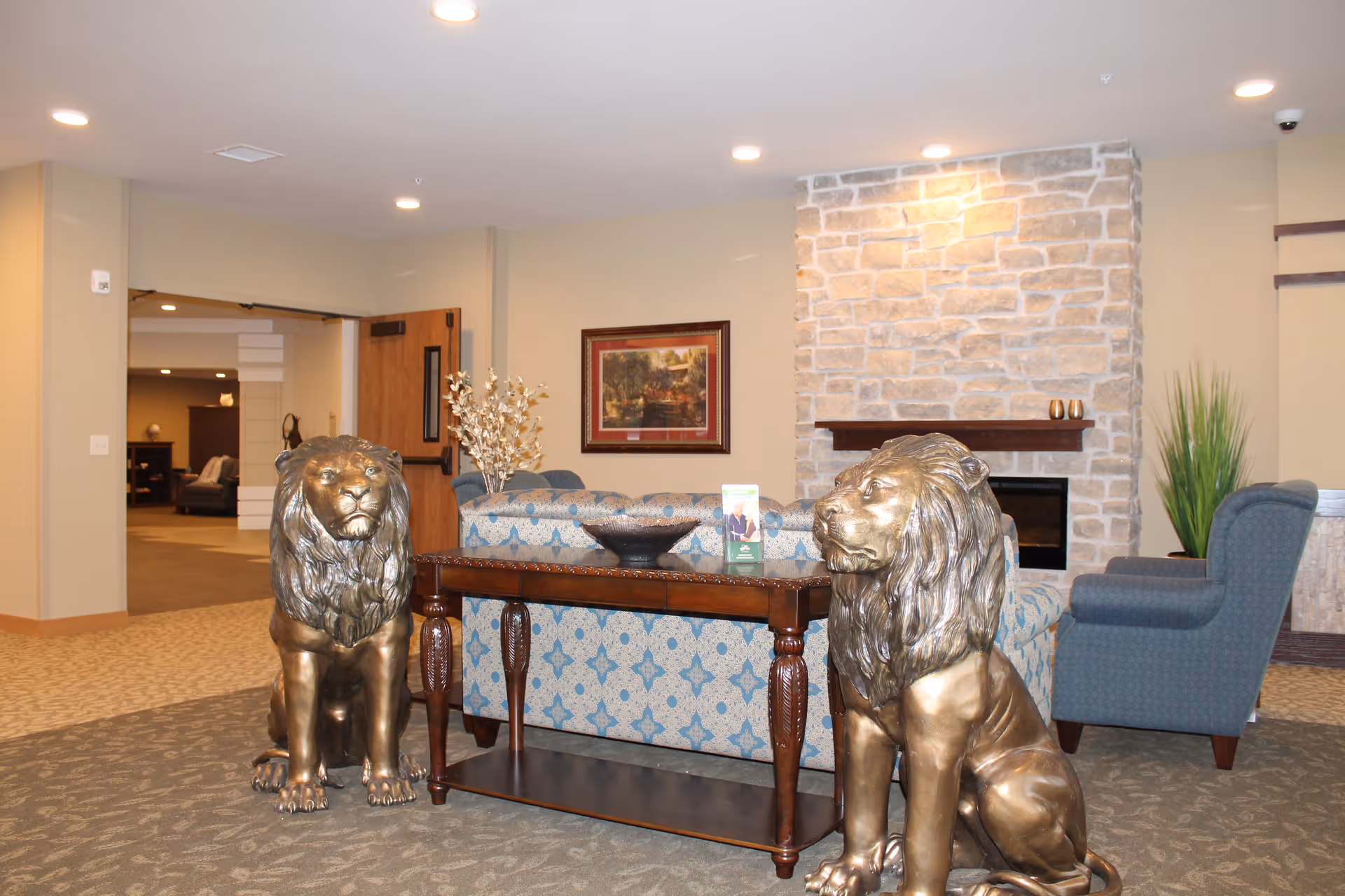 A cozy living room area with a stone fireplace, a patterned blue sofa, a wooden table, two large bronze lion statues, a blue armchair, and a framed painting on the wall.