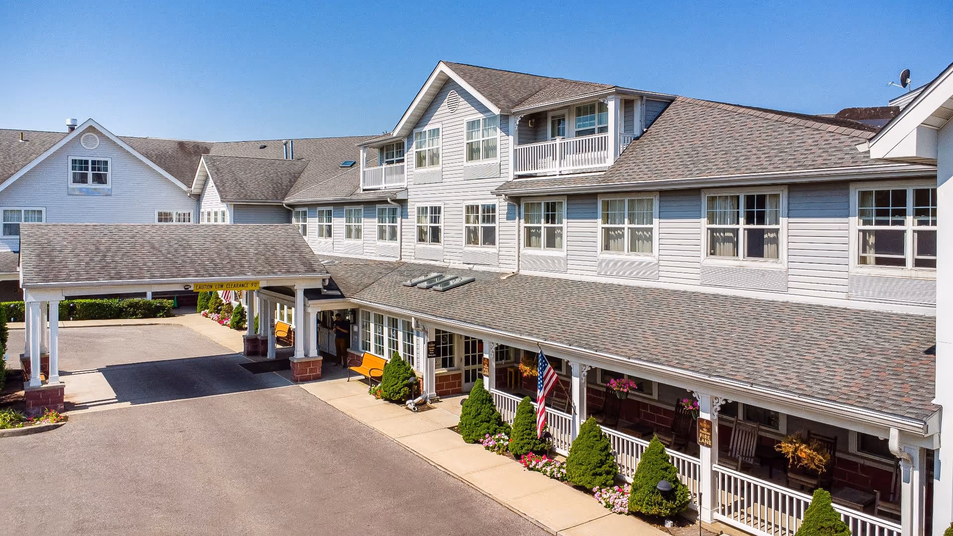 Exterior view of a multi-story senior living facility with a covered entrance, multiple windows, benches, and landscaped bushes and flowers along the sidewalk under a clear blue sky.