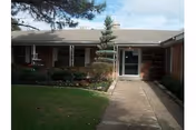 Exterior view of a single-story brick building with a concrete walkway leading to a glass door entrance, surrounded by shrubs and a small tree, under a partly cloudy sky.