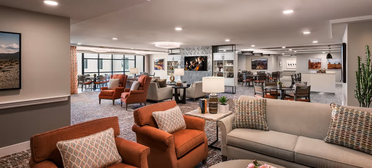 A spacious and well-lit senior living facility common area featuring comfortable seating with beige and rust-colored armchairs and sofas, patterned cushions, side tables with lamps, and a wall-mounted TV. The room has large windows, framed landscape artwork, and multiple dining tables with chairs in the background.
