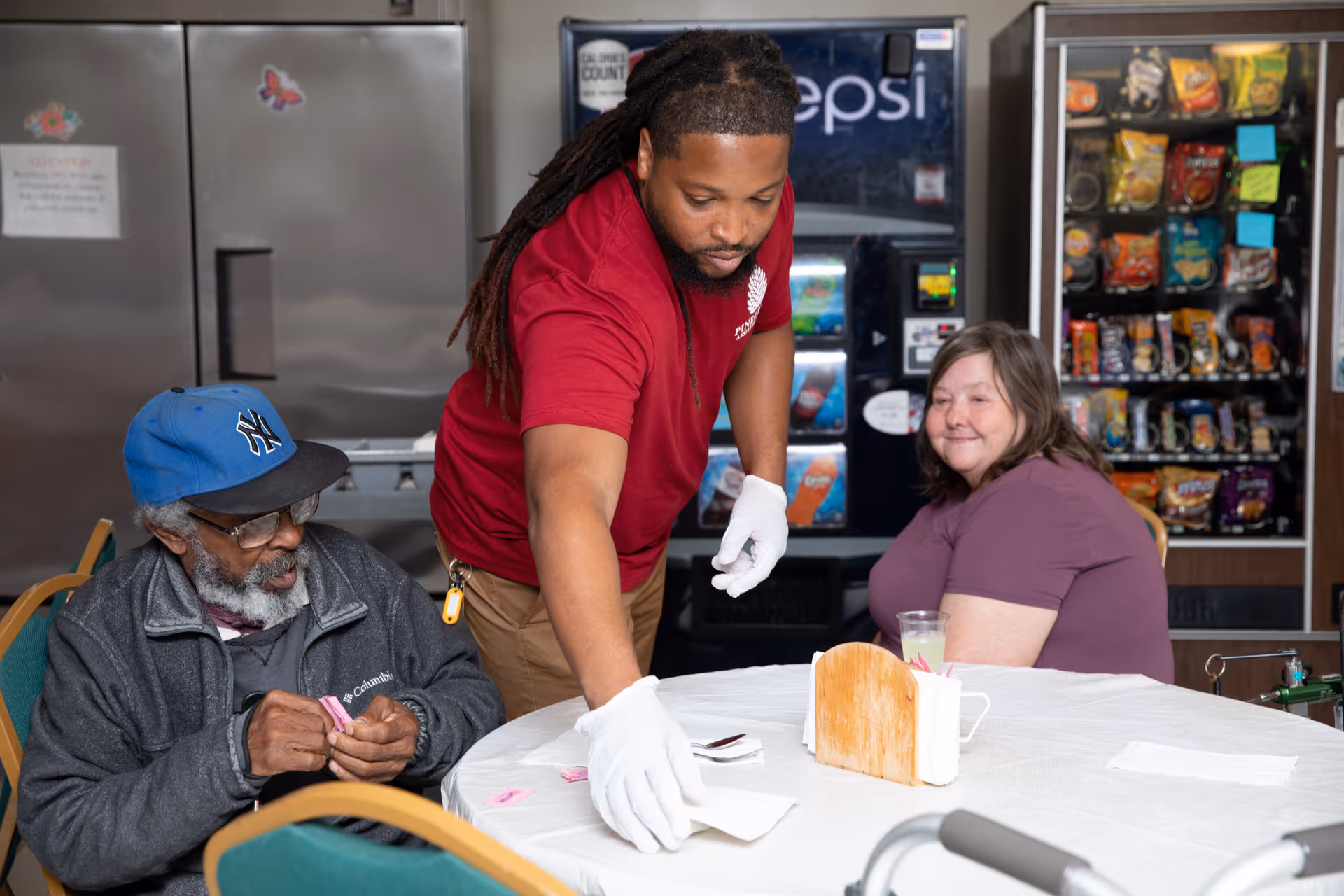 A staff member wearing a red PineCrest Assisted Living shirt and white gloves is placing napkins on a round table covered with a white tablecloth. An elderly man wearing a blue cap and glasses is seated at the table, holding pink tickets, while a woman in a purple shirt sits nearby smiling. Behind them are vending machines stocked with snacks and drinks.