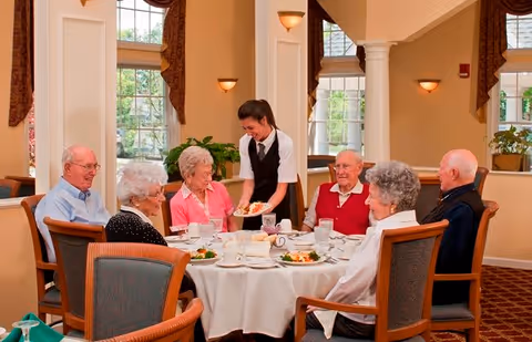 A group of six elderly people sitting around a round dining table with white tablecloth in a well-lit dining room. A waitress is serving food to one of the elderly women. The table is set with plates of food, cups, and glasses. Large windows with curtains and warm lighting fixtures are visible in the background.