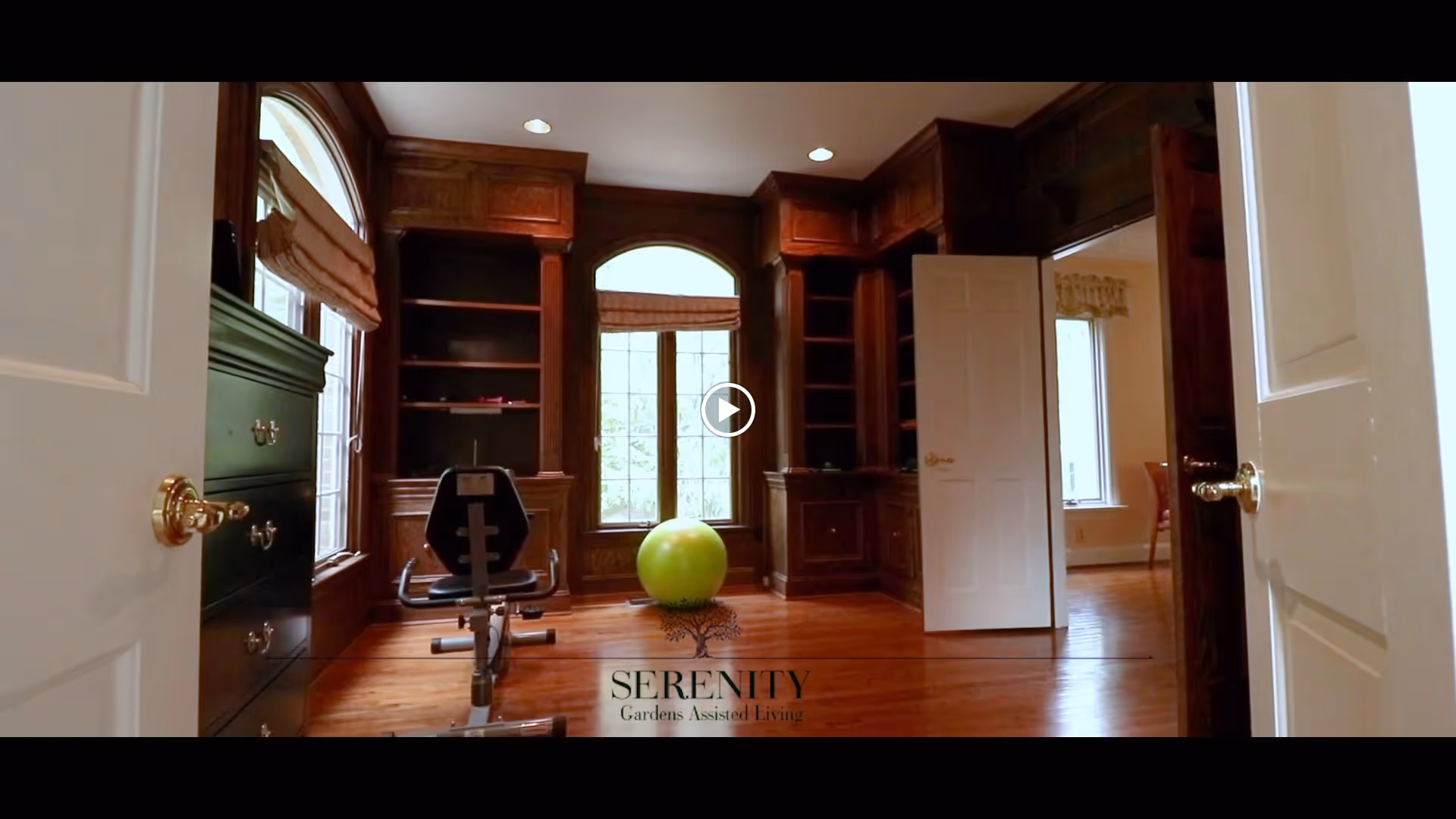Interior room with wooden built-in shelves and cabinets, a large window with an arched top and blinds, a green exercise ball, and a piece of exercise equipment. The room has wooden floors and white doors partially open to adjacent rooms.