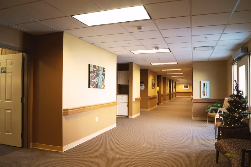 Carpeted hallway inside a care facility with handrails, wall art, seating and a small decorated Christmas tree.