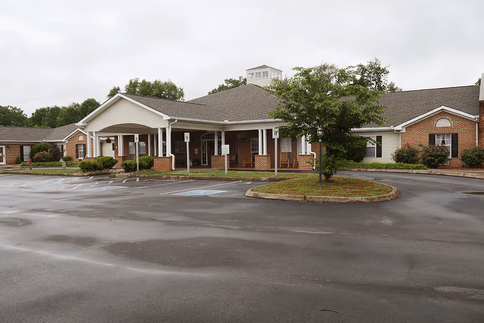 Exterior view of Brookdale Browns Creek senior living facility showing a single-story brick building with a covered entrance, several windows, and a small tree in a landscaped island in the parking lot on a cloudy day.