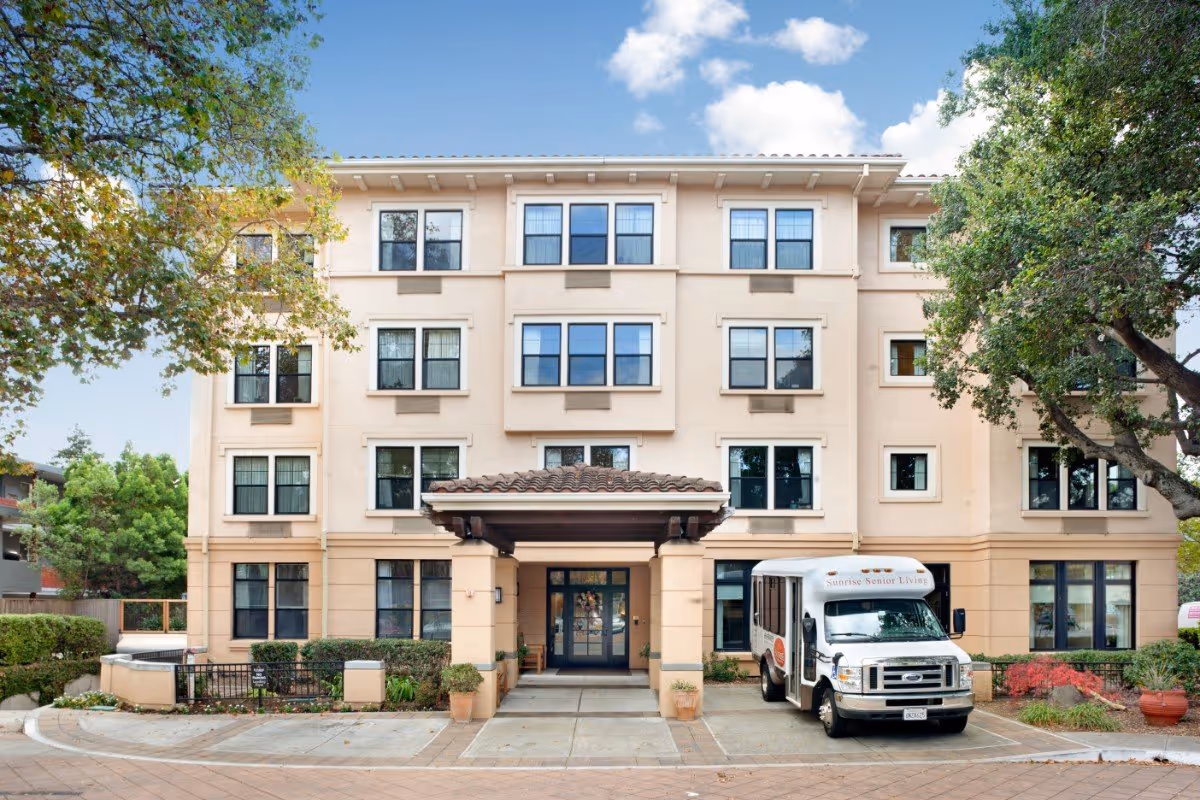 Four-story beige senior living building front with a covered entrance and a Sunrise Senior Living shuttle van parked in front, framed by trees.