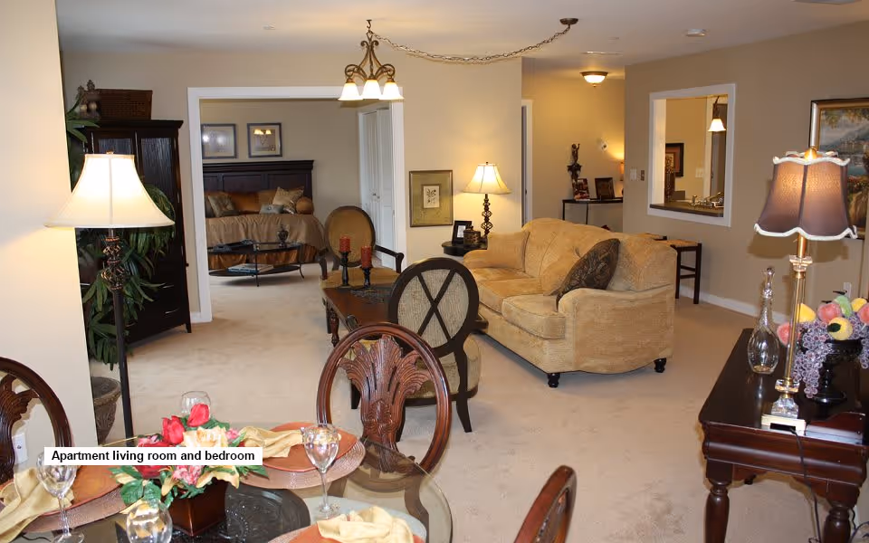View of an apartment living room and bedroom area with beige carpet and walls. The living room features a beige sofa, two chairs, a coffee table, lamps, and decorative items. The bedroom is visible through an open doorway, showing a bed with pillows and framed pictures on the wall. A dining table with chairs and place settings is in the foreground.