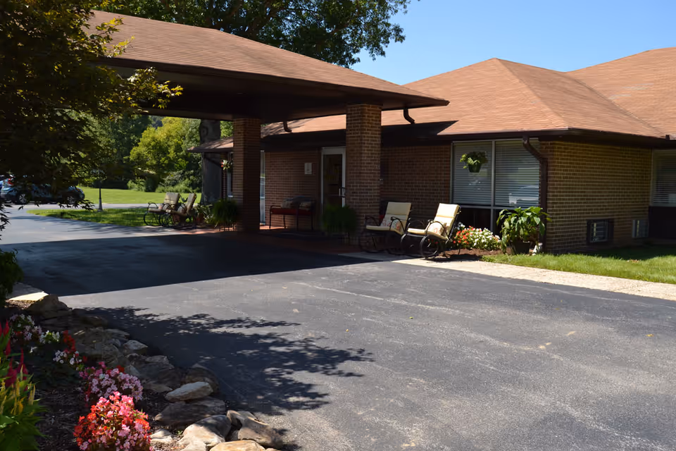 Front entrance of a brick senior living facility with a covered driveway, outdoor chairs, and flowerbeds.