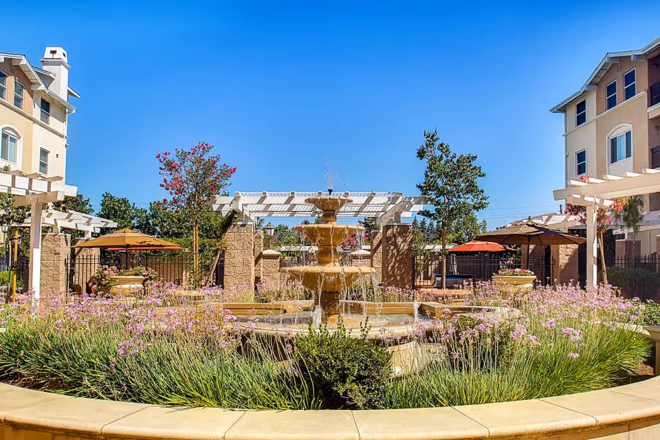 Outdoor courtyard area at Heritage Estates featuring a central tiered water fountain surrounded by lush greenery and purple flowers. There are pergolas, patio umbrellas, and seating areas with buildings visible in the background under a clear blue sky.