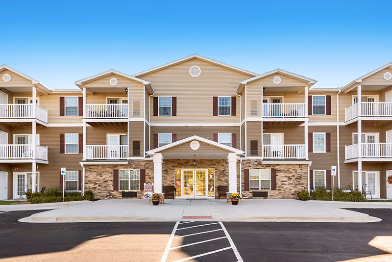Front exterior view of a three-story senior living facility building with beige siding, stone accents, multiple balconies, and a covered entrance with white columns. There is a parking area and a clear blue sky above.