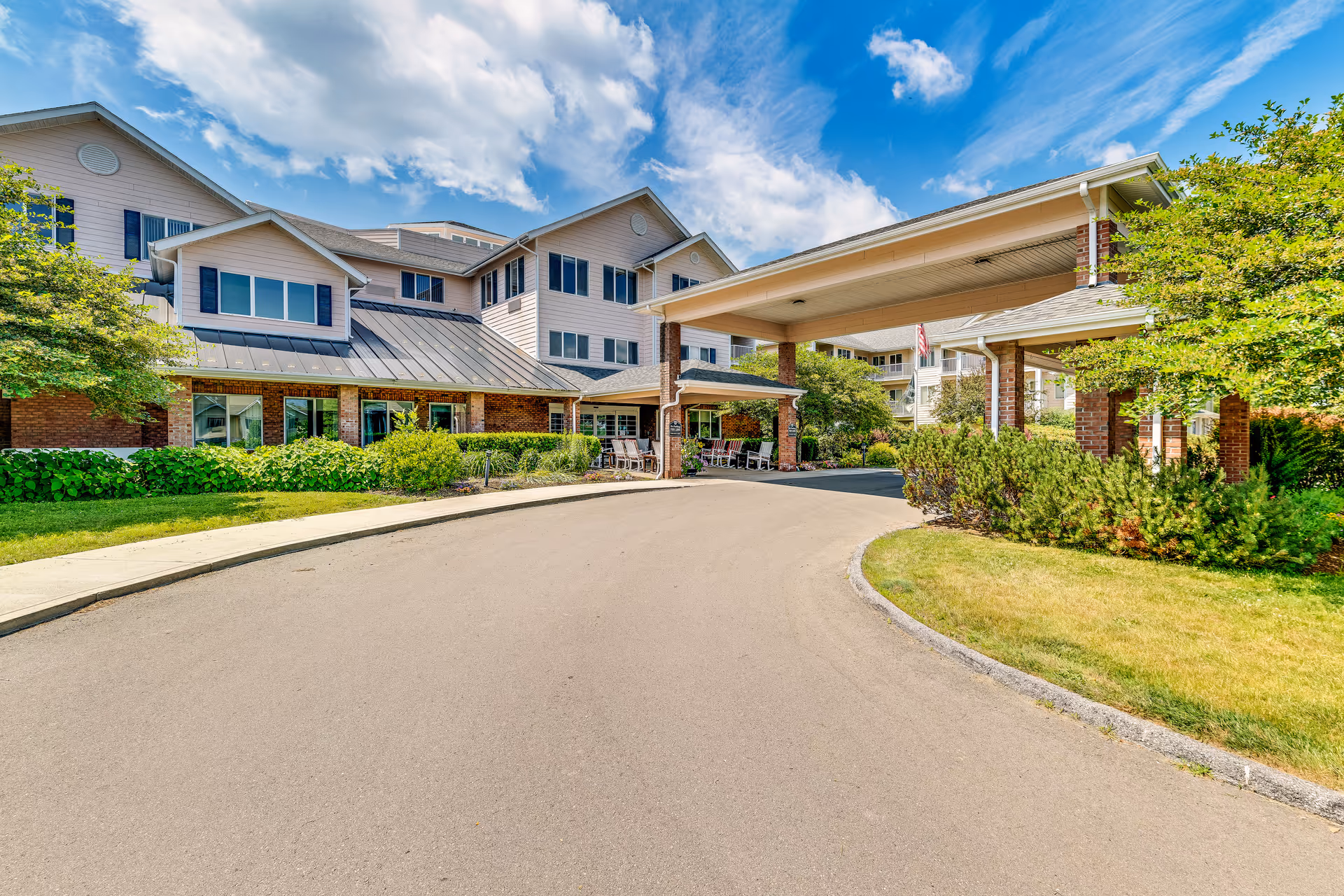 Exterior view of Solstice Senior Living at Bangor showing a large multi-story building with a covered entrance driveway, surrounded by green bushes and trees under a partly cloudy blue sky.