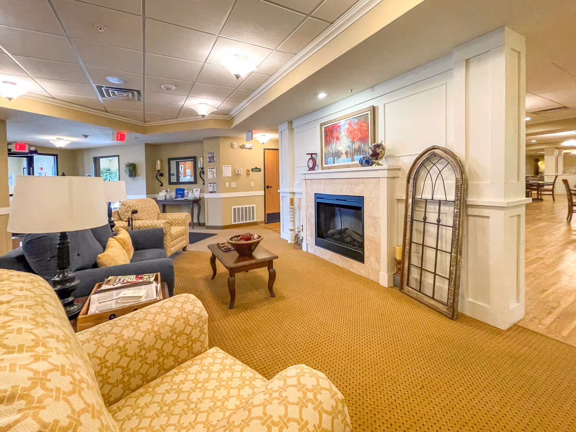 Cozy common room with upholstered armchairs, a coffee table and a decorative fireplace in a senior living facility.