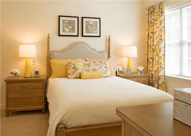 A cozy bedroom with a wooden bed frame and a beige upholstered headboard. The bed is made with a cream-colored quilt and decorated with yellow and floral-patterned pillows. On each side of the bed are wooden nightstands with yellow lamps, small flower arrangements, and framed photos. Two framed floral artworks hang above the headboard. A window with yellow floral curtains allows natural light into the room.