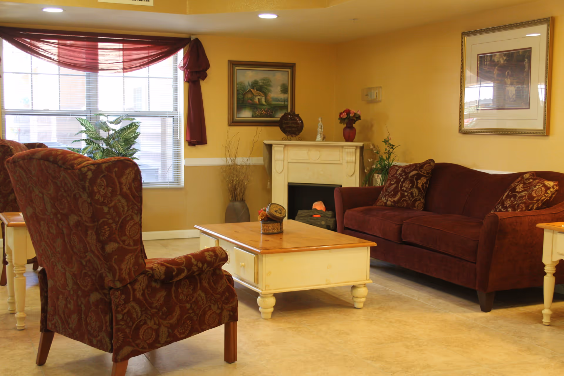 A cozy living room with a maroon patterned armchair and a matching maroon sofa with decorative pillows. A light wooden coffee table sits in the center on a tiled floor. The room features a white fireplace with decorative items on the mantel, framed artwork on the walls, and a window with blinds and a maroon curtain swag. There are also some plants and a vase with dried branches in the corner.