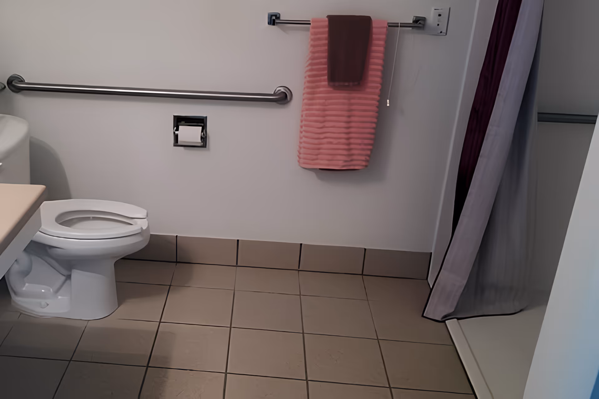 A bathroom with a white toilet, beige tiled floor, a grab bar on the wall, a toilet paper holder, and two towels hanging on a towel rack above the toilet paper holder. There is a shower area with a curtain partially visible on the right side.
