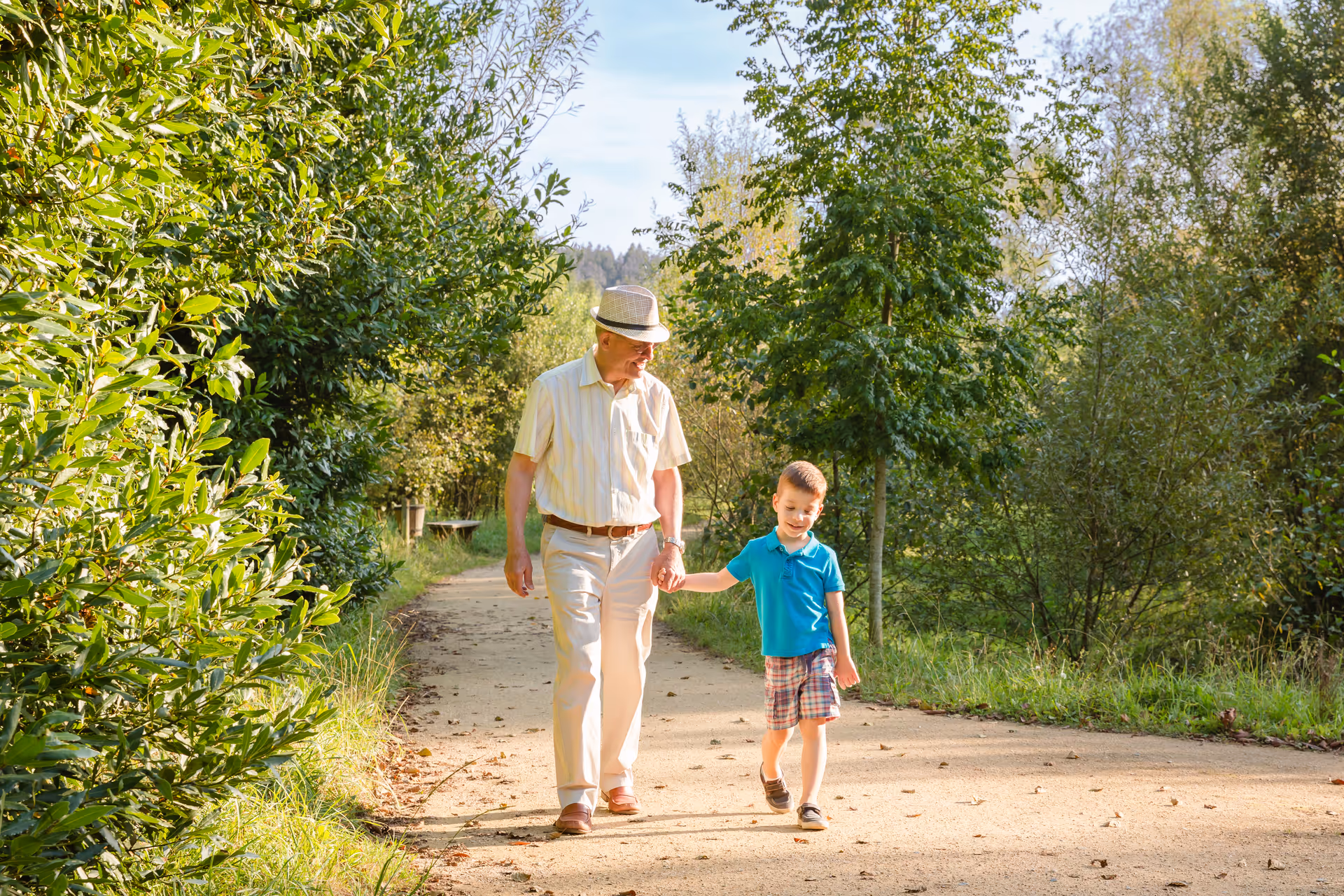 An elderly man wearing a hat and light-colored clothes is walking hand in hand with a young boy in a blue shirt and plaid shorts along a sunlit path surrounded by green trees and bushes.