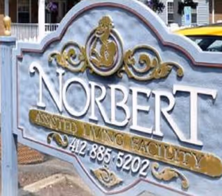 Close-up of a decorative sign for Norbert Assisted Living Facility with a phone number 412-885-5202, featuring ornate gold and white lettering on a blue background.
