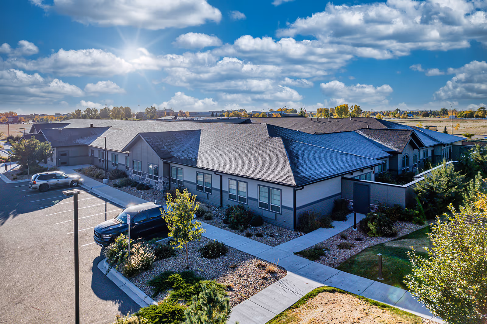 Aerial view of a single-story building with a dark roof and light-colored walls, surrounded by landscaped greenery and a parking lot with a few vehicles. The sky is partly cloudy with the sun shining brightly.