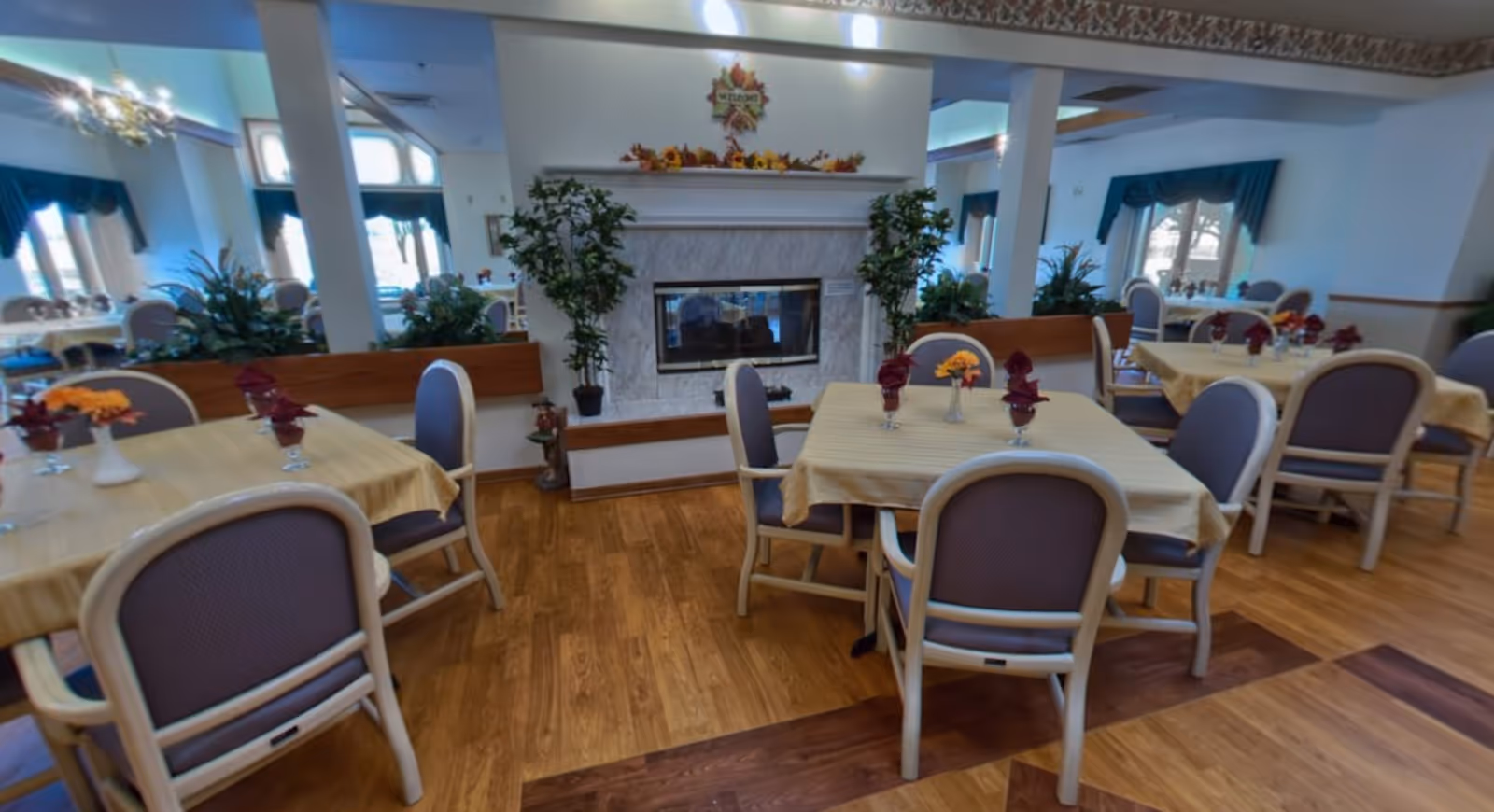 A dining room with several tables covered with yellow tablecloths, each set with chairs and small floral centerpieces. The room features a fireplace with a decorative mantle adorned with autumn-themed decorations and plants on either side. Large windows with blue curtains allow natural light to fill the space, and the floor is made of wood.