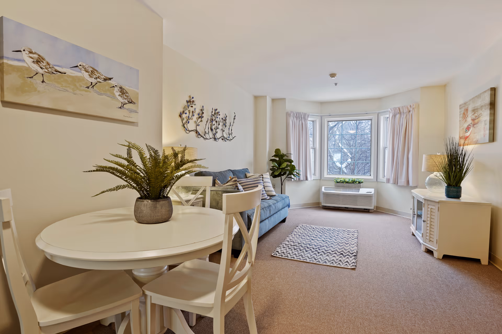A cozy living room area in a senior living facility with a round white dining table and chairs on the left, a blue sofa with striped pillows against the wall, a small patterned rug on the carpeted floor, a bay window with curtains, and a white cabinet with a lamp and plant on the right. Wall decorations include a painting of birds and a metal wall art piece.
