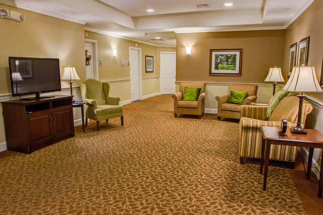 Well-lit senior living common room with upholstered chairs and sofas, a TV on a cabinet, side tables with lamps, and framed artwork on the walls.