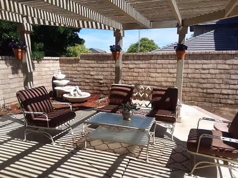 Outdoor patio area with a pergola casting striped shadows over cushioned chairs and a glass-top coffee table. There are potted plants hanging from the pergola and a small water fountain against a brick wall in the background.