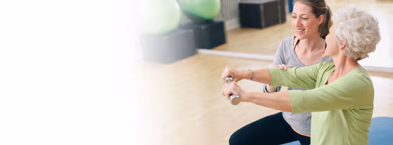 A senior woman in a green top is exercising with a fitness bar while a younger woman in a gray top assists and encourages her in a bright indoor space with exercise balls in the background.