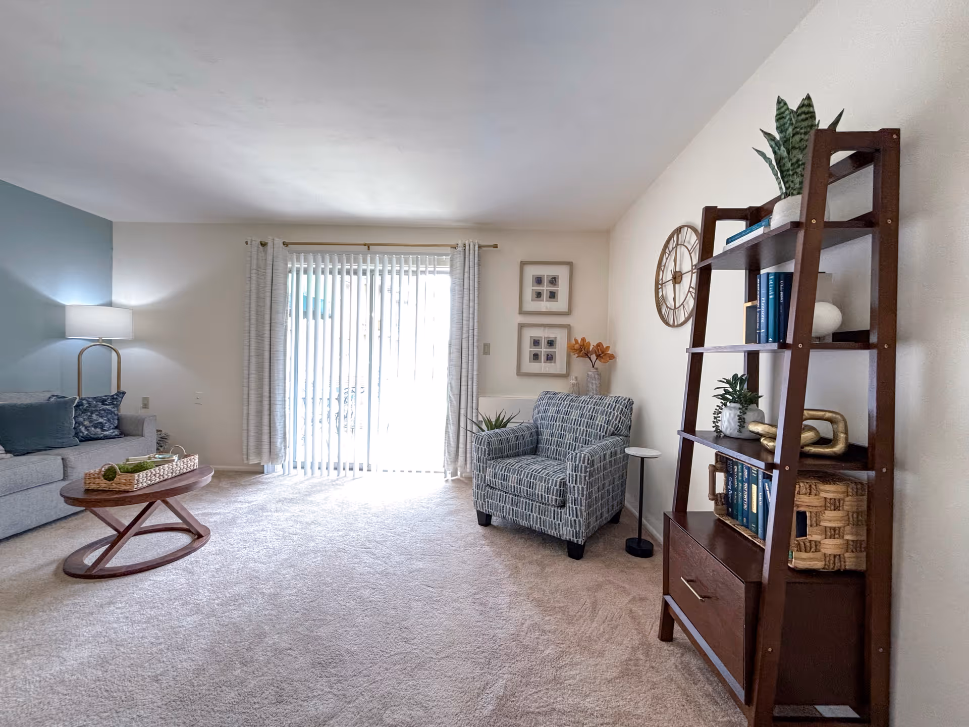 A bright living room with beige carpet, a gray sofa with blue cushions, a wooden coffee table with a decorative tray, a patterned armchair, a tall wooden bookshelf with books and plants, a wall clock, and vertical blinds covering a sliding glass door.