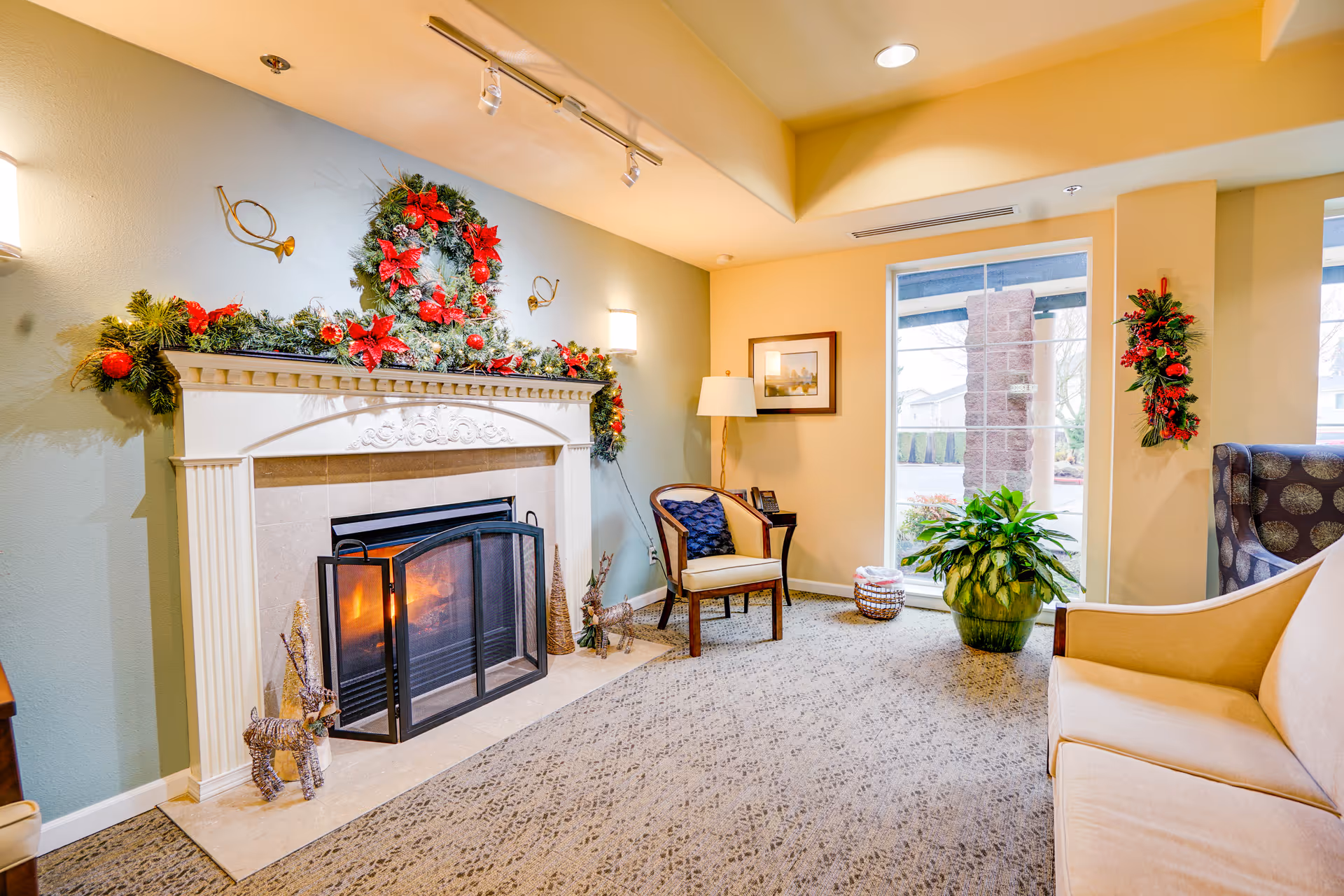 Cozy lounge area with a lit fireplace, a decorated mantel with holiday wreaths, seating, and a window.