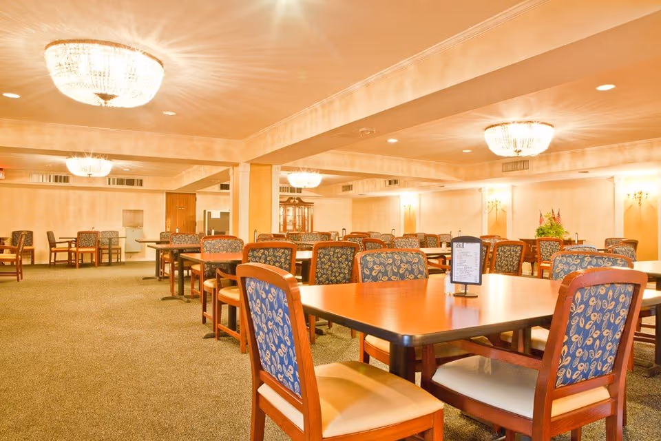 A spacious dining room with multiple wooden tables and cushioned chairs featuring a blue patterned fabric. The room is well-lit with large, elegant chandeliers on the ceiling and wall sconces providing additional light. The carpeted floor and neutral-colored walls create a warm and inviting atmosphere.
